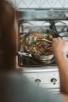 Person cooking colorful stir-fry on gas stove, featuring mixed vegetables and steam.