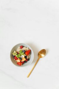 A colorful breakfast bowl with quinoa, fresh fruits, and a golden spoon on a white surface.